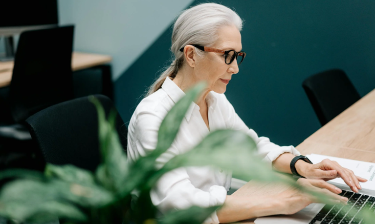 A retirement-age older woman with silver hair works on her laptop at her office table