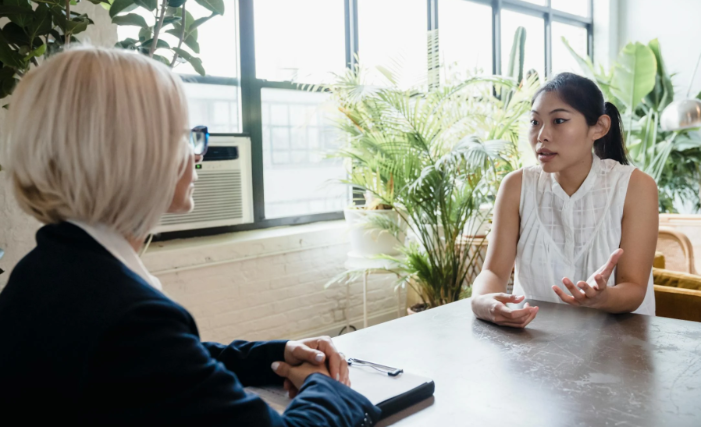 a younger employee is at a desk speaking with her more senior colleague at a mentoring session at work