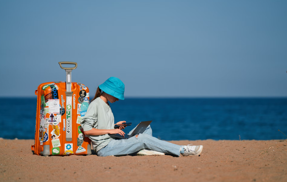 digital nomad leans on her luggage while working on her laptop at the beach