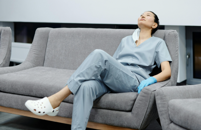 nurse in scrubs and gloves is seated on a couch, her eyes closed, tired from the stressful day at work