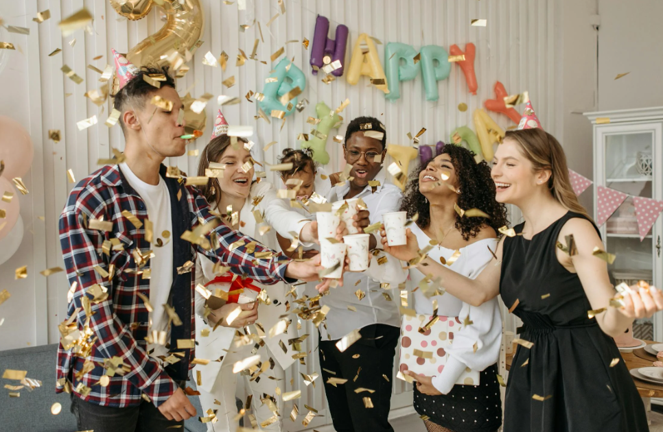 a group of people celebrating a birthday with drinks, party hats, and confetti 