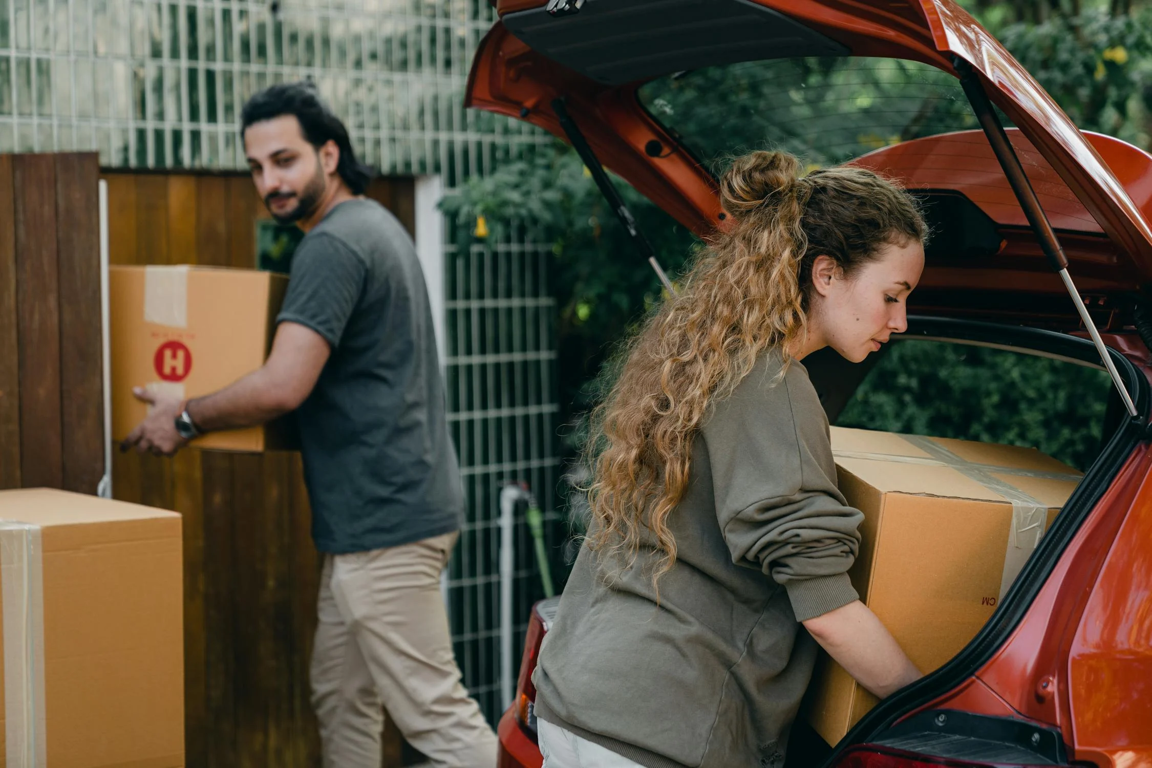 a millennial couple lifting boxes of their belongings from a red car in front of their new home