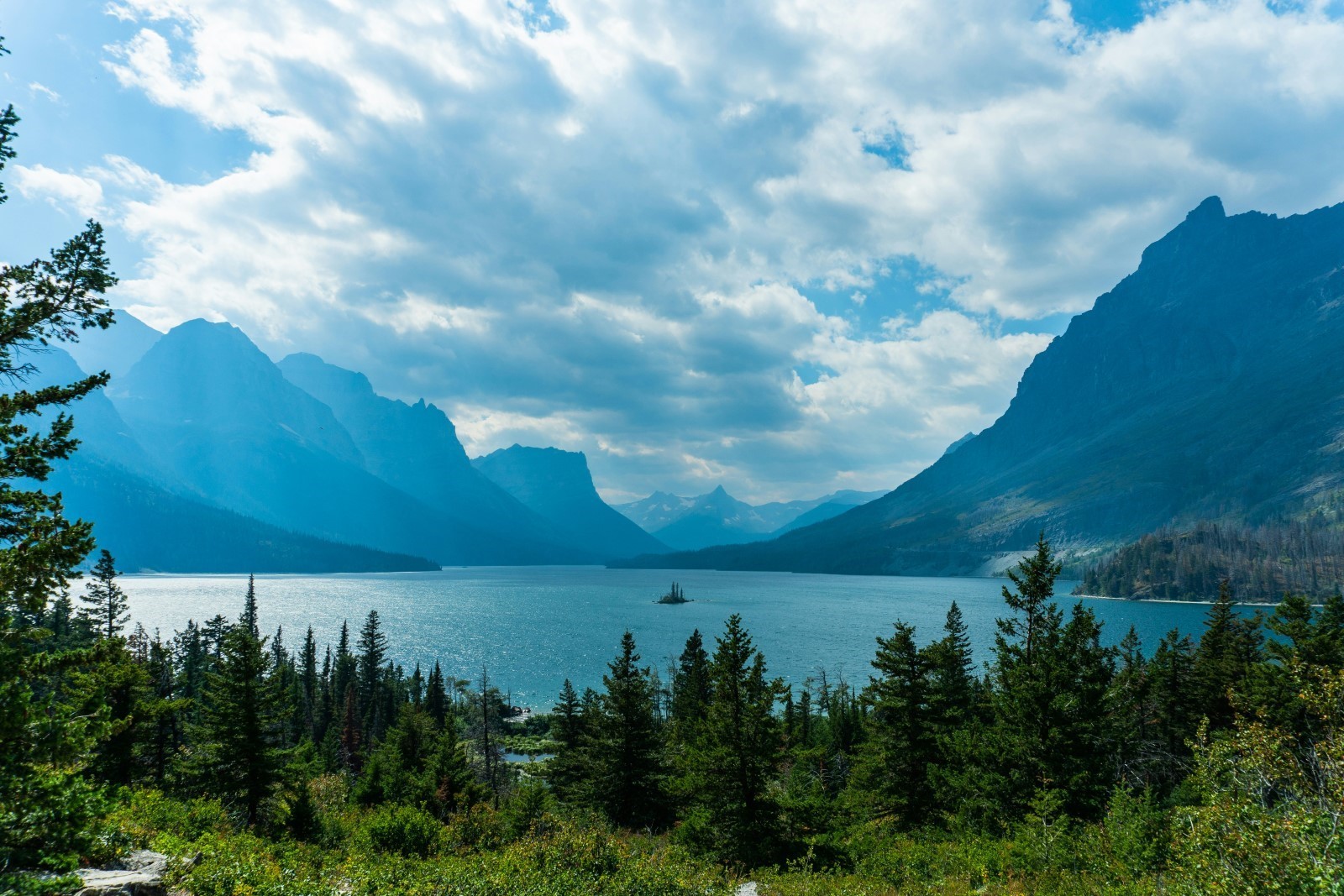 Lake Josephine in Glacier National Park in Montana on a clear day, with trees in the foreground and blue mountains beyond the lake