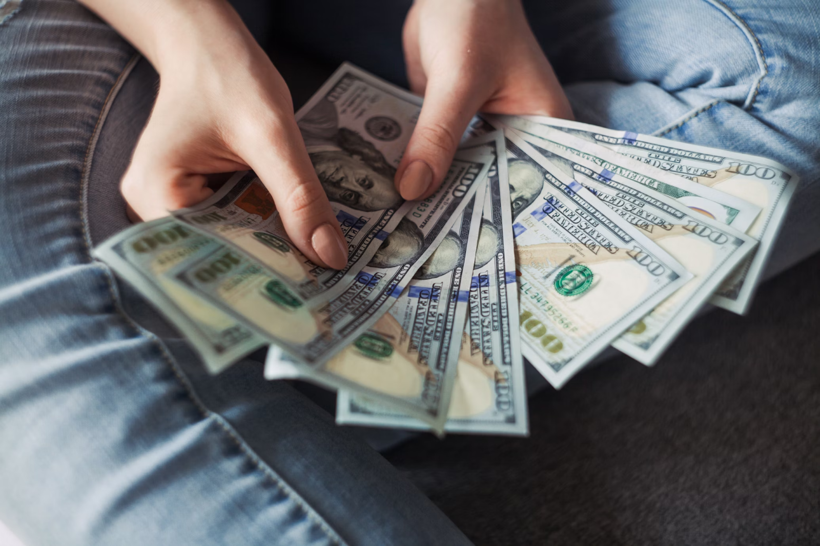 a woman wearing denim pants counts American dollar bills while sitting on black carpeted floor