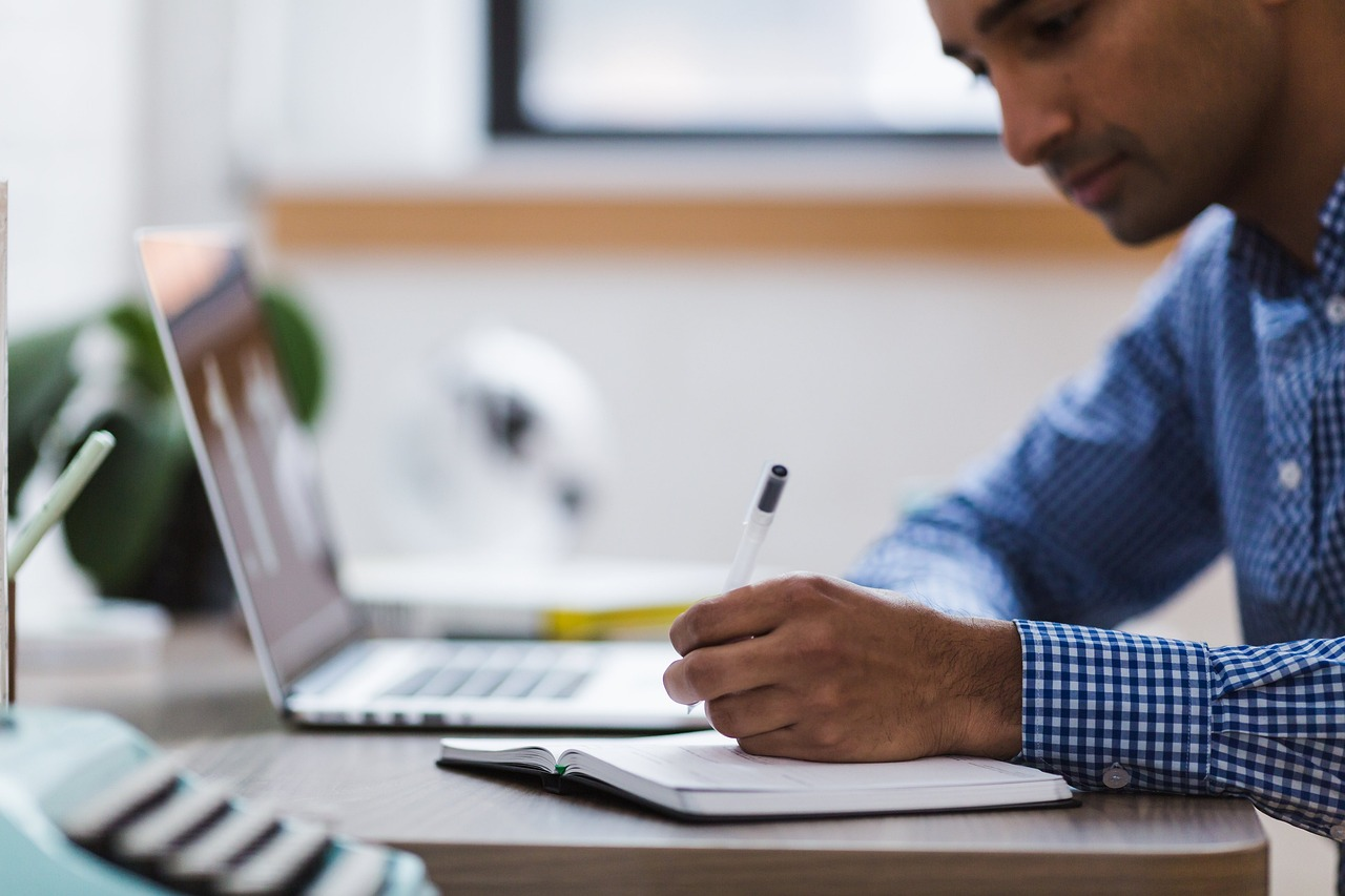 an aspiring mortgage specialist is completing his educational requirements using a laptop while also writing on a notebook