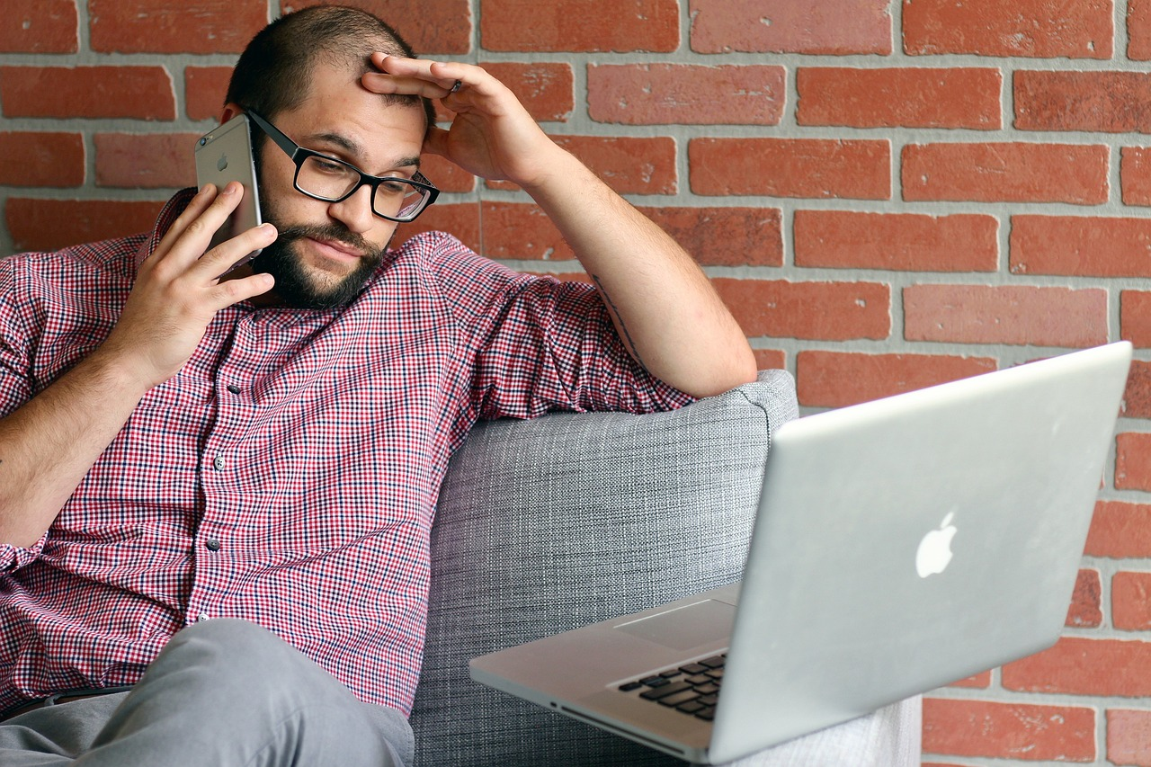  a bearded man wearing red checkered shirt holding his phone and looking at laptop while stressing over his mortgage bills