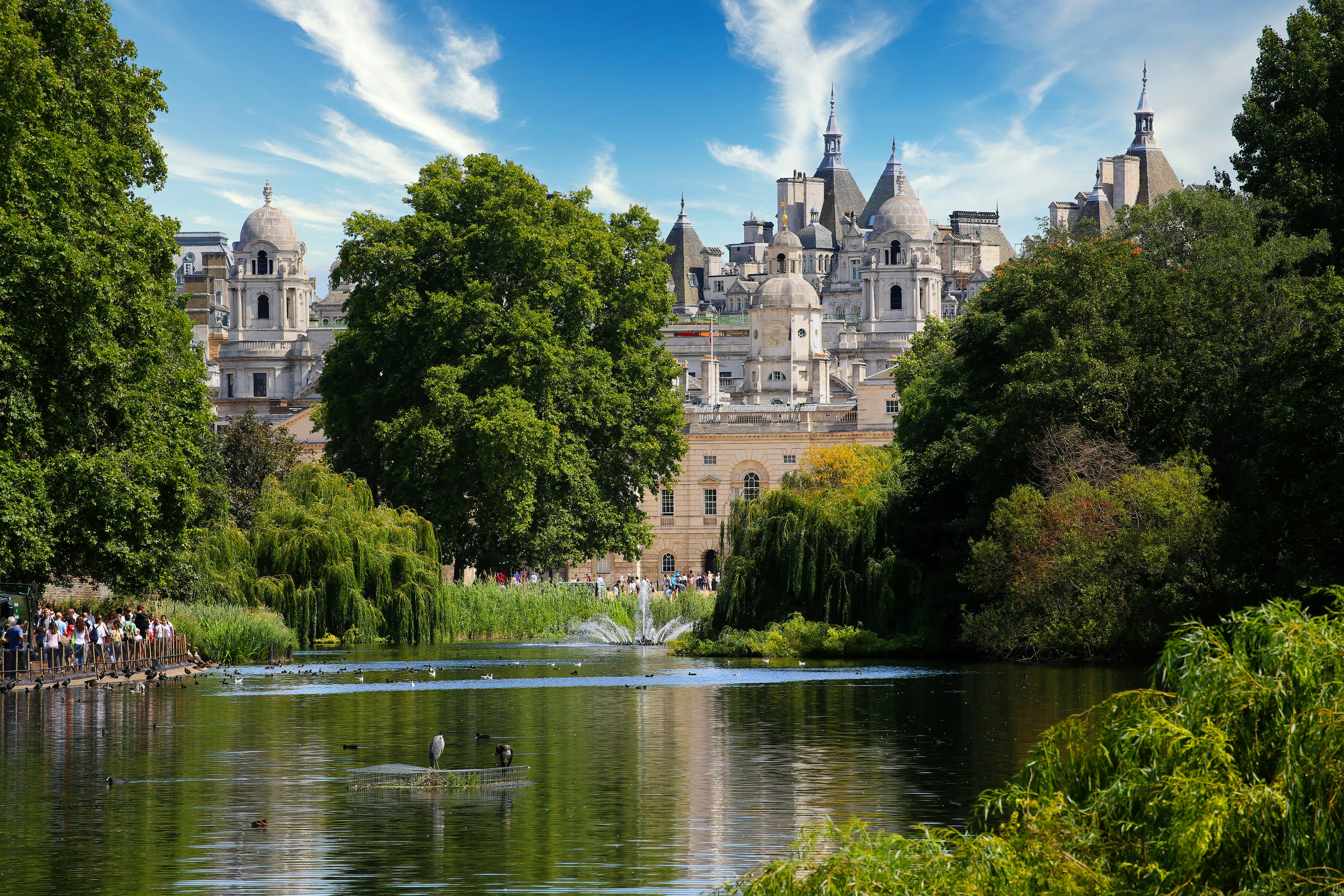 people enjoying a sunny day in Hyde Park, surrounded by lush greenery and a serene lake with medieval buildings in the background