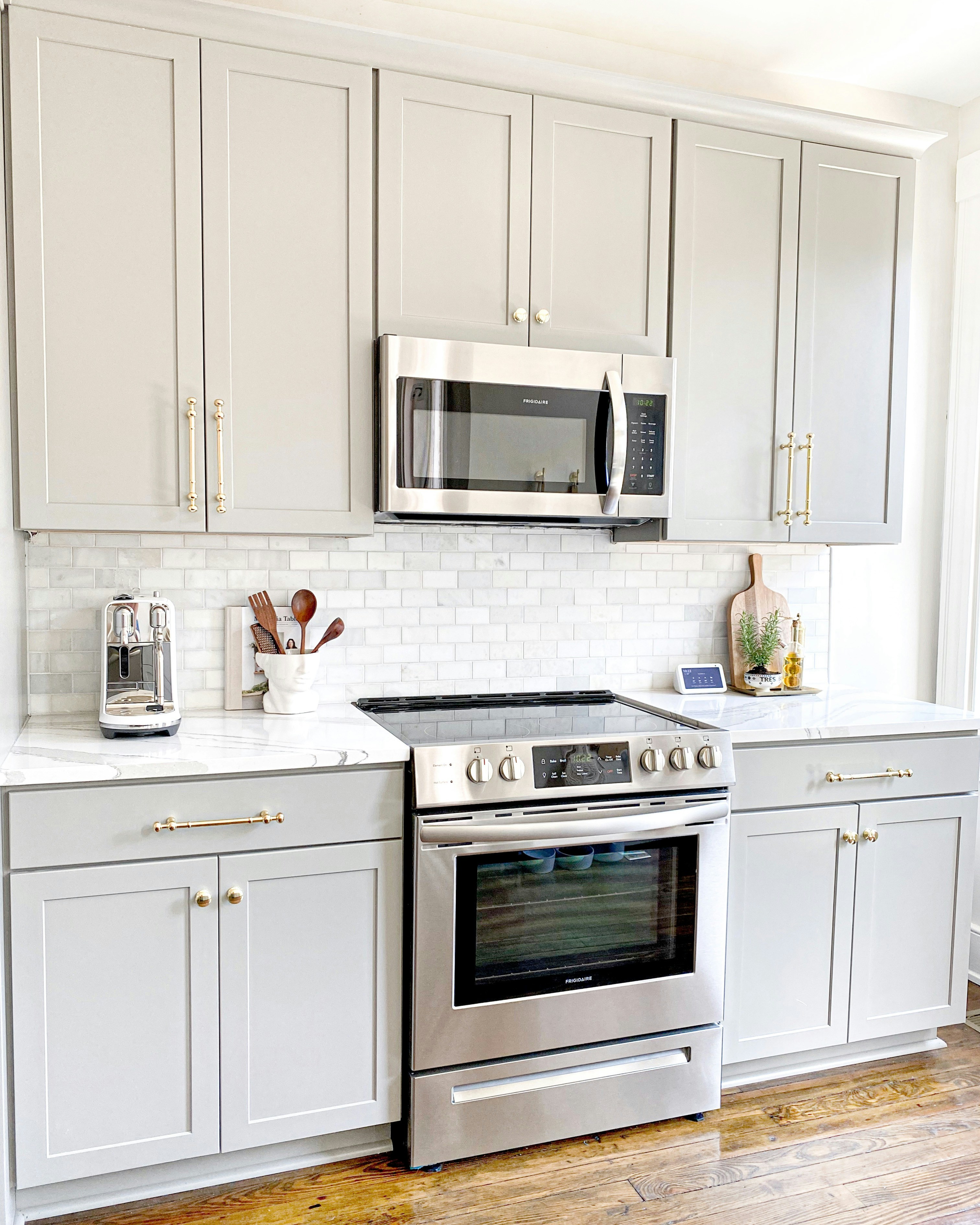 an all-white kitchen with shiny knobs and drawer pulls, modern cooktop stove and matching microwave above it, and a white brick backsplash; and below, solid hardwood floors